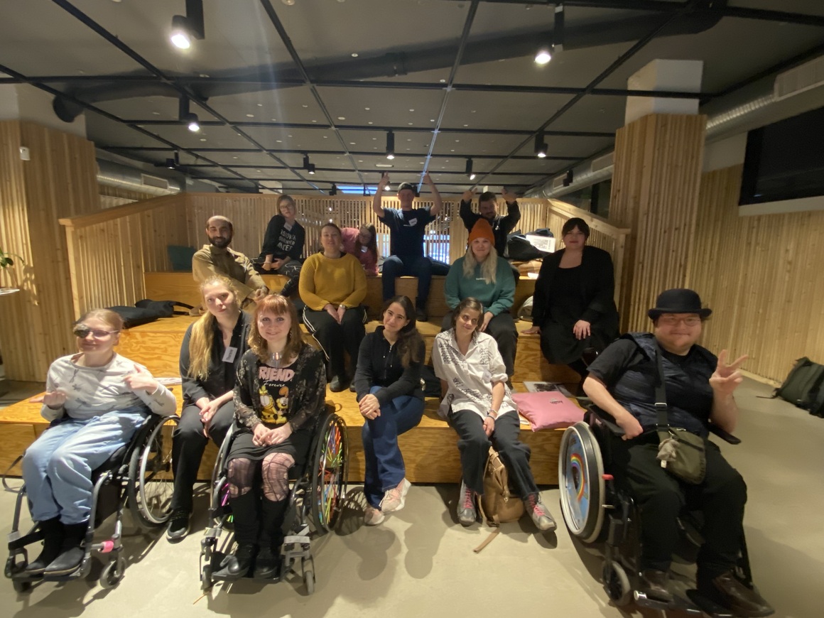 Gruppbild från konstnärsträffen. 13 deltagare. sitter i och framför en läktare i en föreläsningssal.