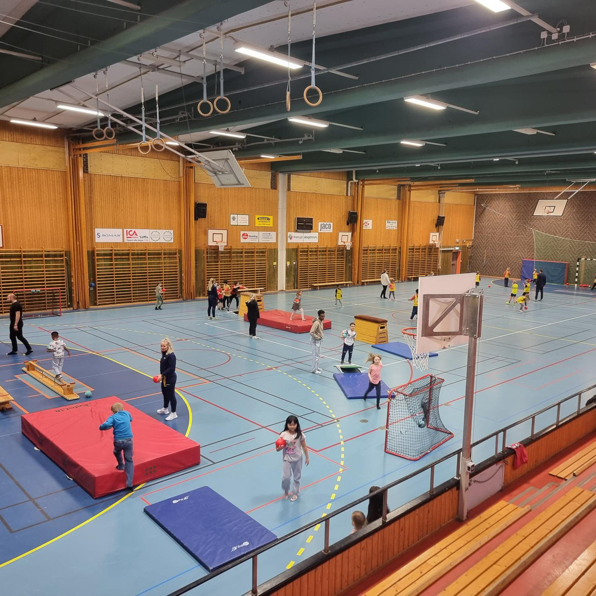 ett bild med barn i idrottshall som springer runt vid olika stationer med mattor och bollar