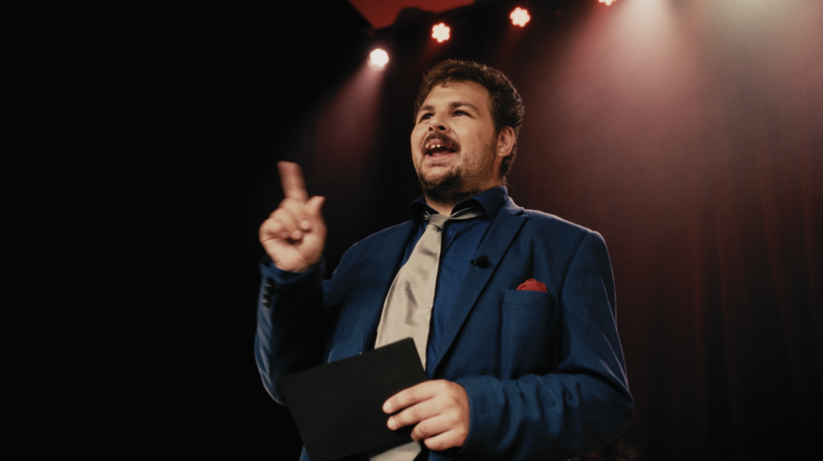 En man i blå kostym och slips står på en scen och pratar.